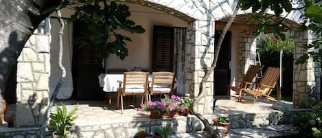 A stone building with a covered patio, wooden chairs, and potted plants.
