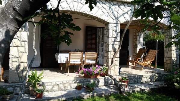 A stone building with a covered patio, wooden chairs, and potted plants.