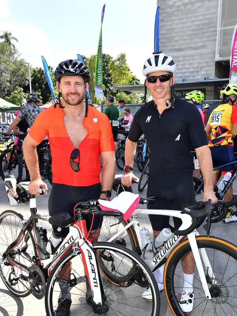 Zwei Radfahrer mit Fahrrädern, einer trägt ein rotes Hemd und der andere Schwarz, stehen in einem belebten Bereich, im Hintergrund weitere Radfahrer und Flaggen.