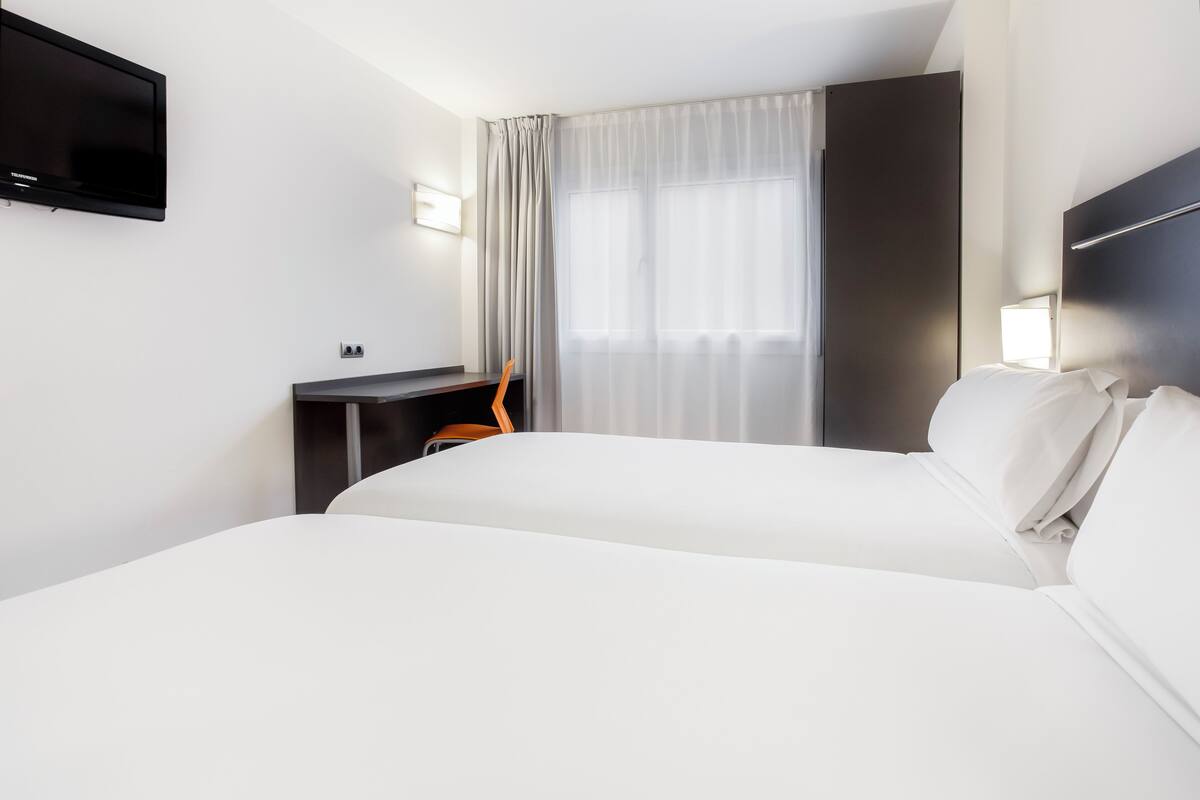 Habitación de hotel con dos camas, un escritorio, una silla, un televisor y una ventana con cortinas.