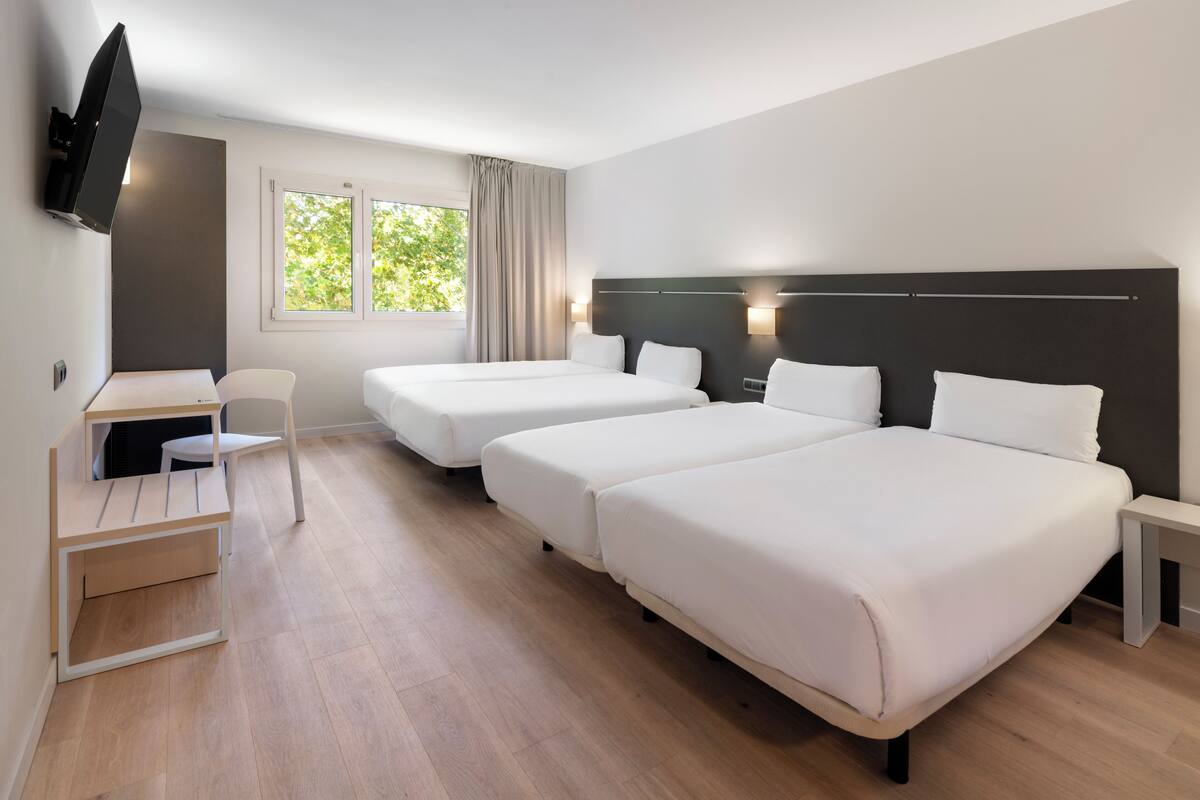 Una habitación de hotel con dos camas individuales, un escritorio con silla, un televisor montado en la pared y una ventana con cortinas.