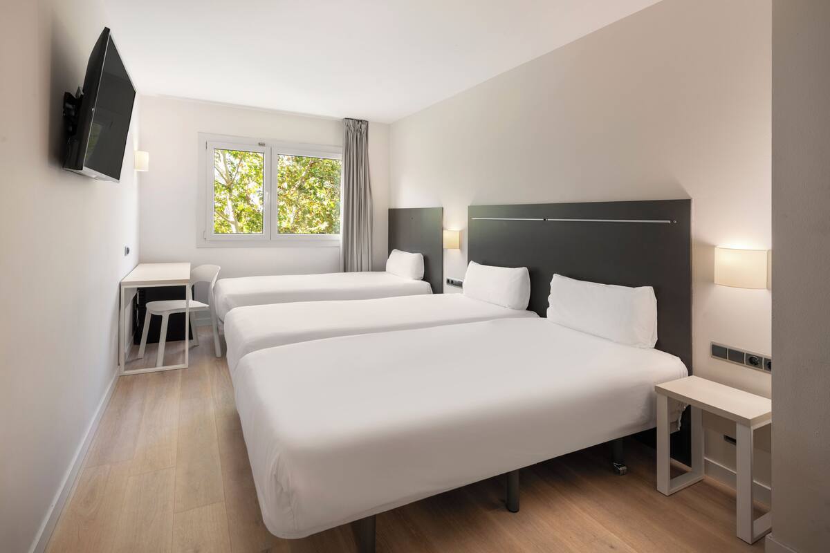 Habitación de hotel con tres camas individuales, un escritorio y una televisión montada en la pared.