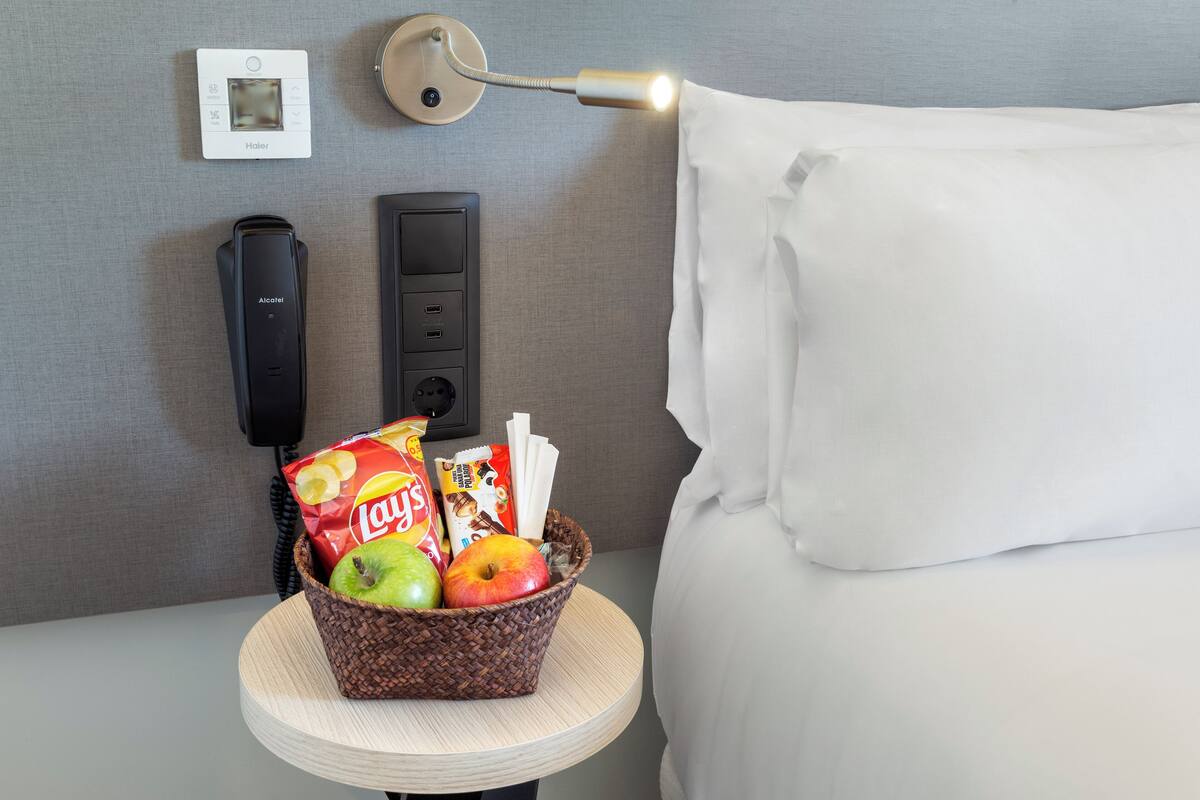 Habitación de hotel con cama, mesita de noche con una cesta de aperitivos, lámpara, teléfono y termostato.