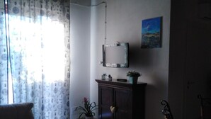 A living room with a sofa, a cabinet, a chair, a television on the wall, and a window with curtains. - STUDIO APARTMENT WITH GARDEN VIEW (Scicli)