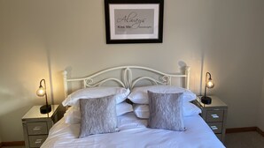 Una cama bien tendida con sábanas blancas y dos almohadas grises, flanqueada por mesitas de noche con lámparas, en una habitación con un cartel enmarcado en la pared.