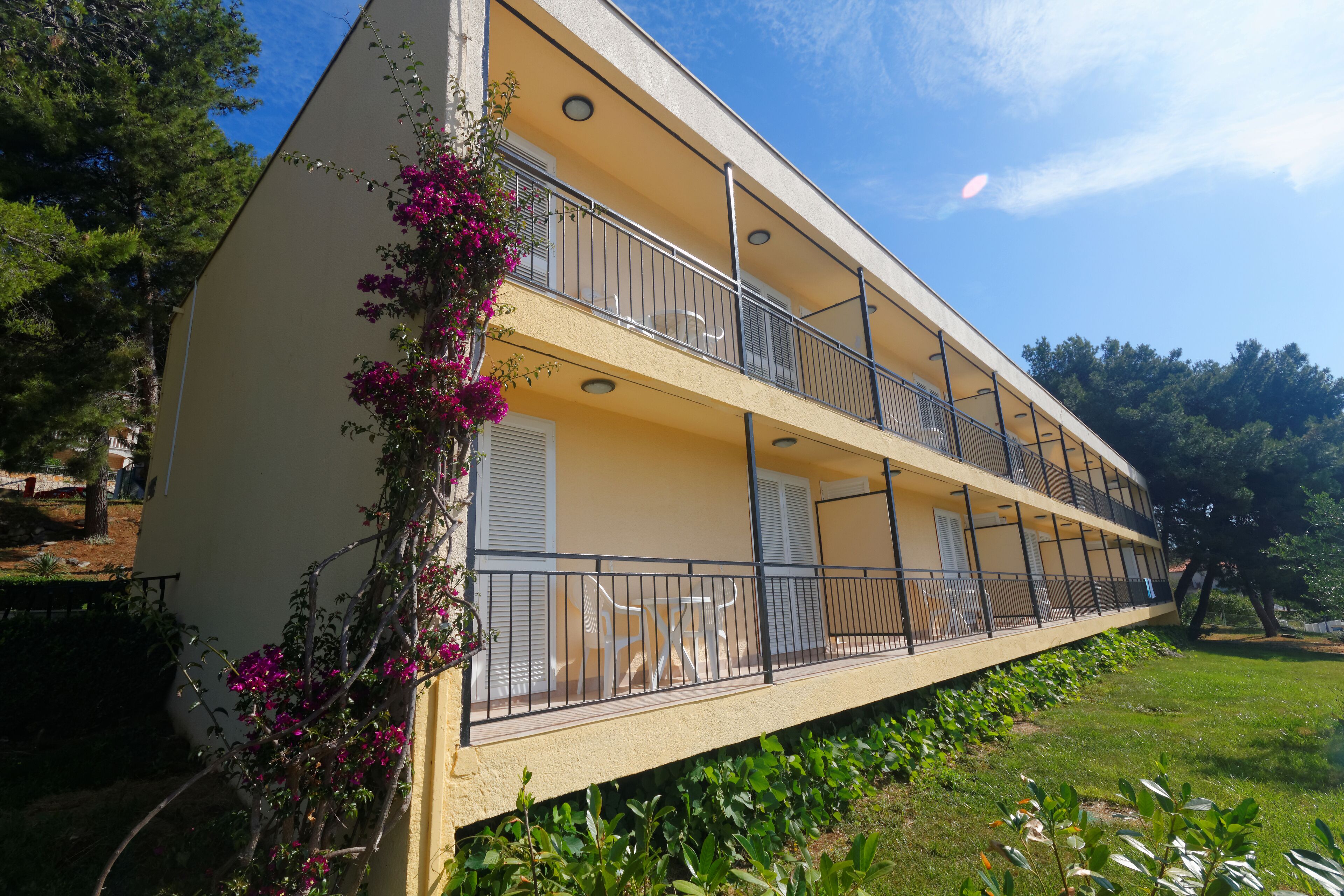 A two-story building with balconies, a garden area, and a clear sky.