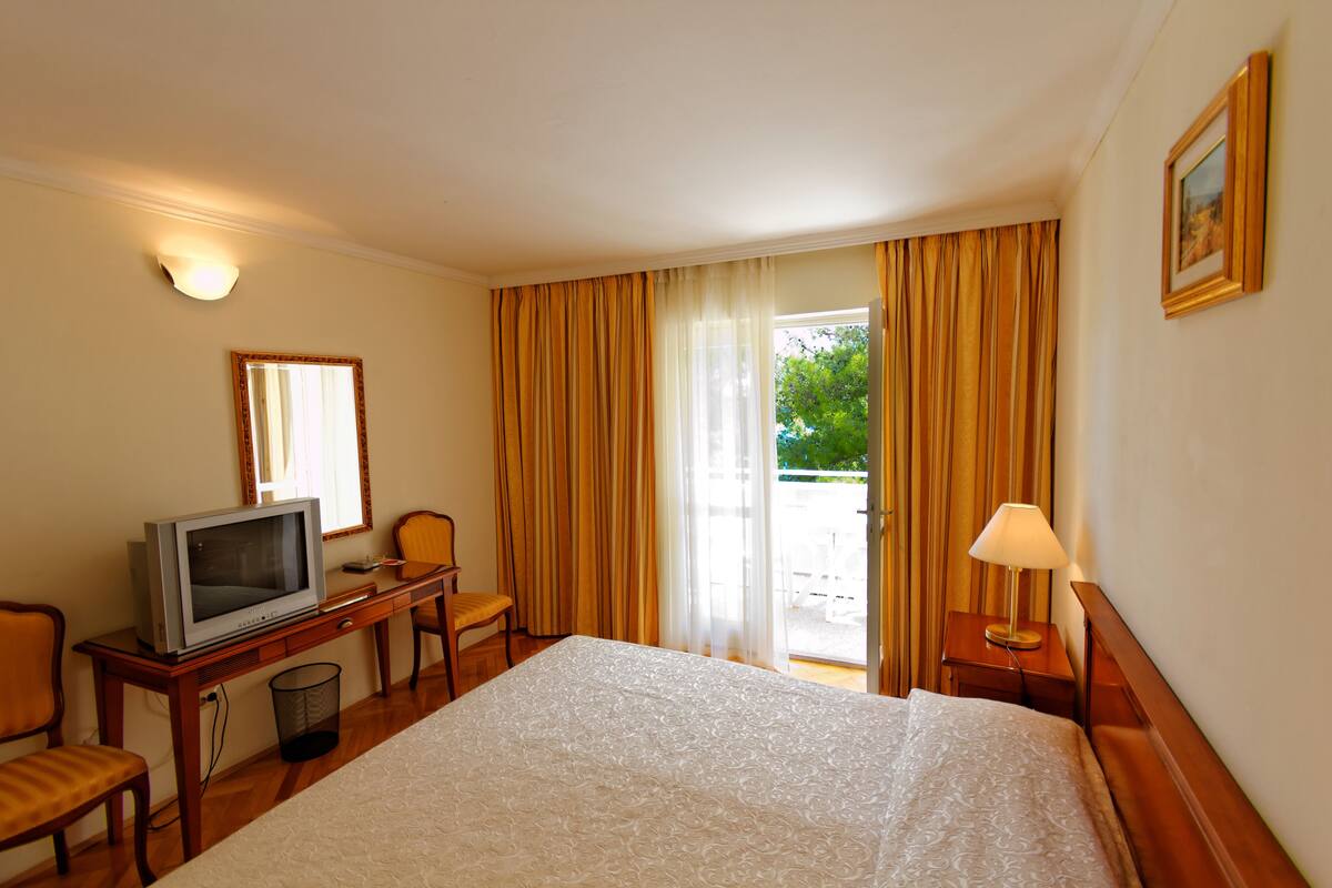 A hotel room with a bed, a desk with a television, a chair, a lamp, and a window with curtains.