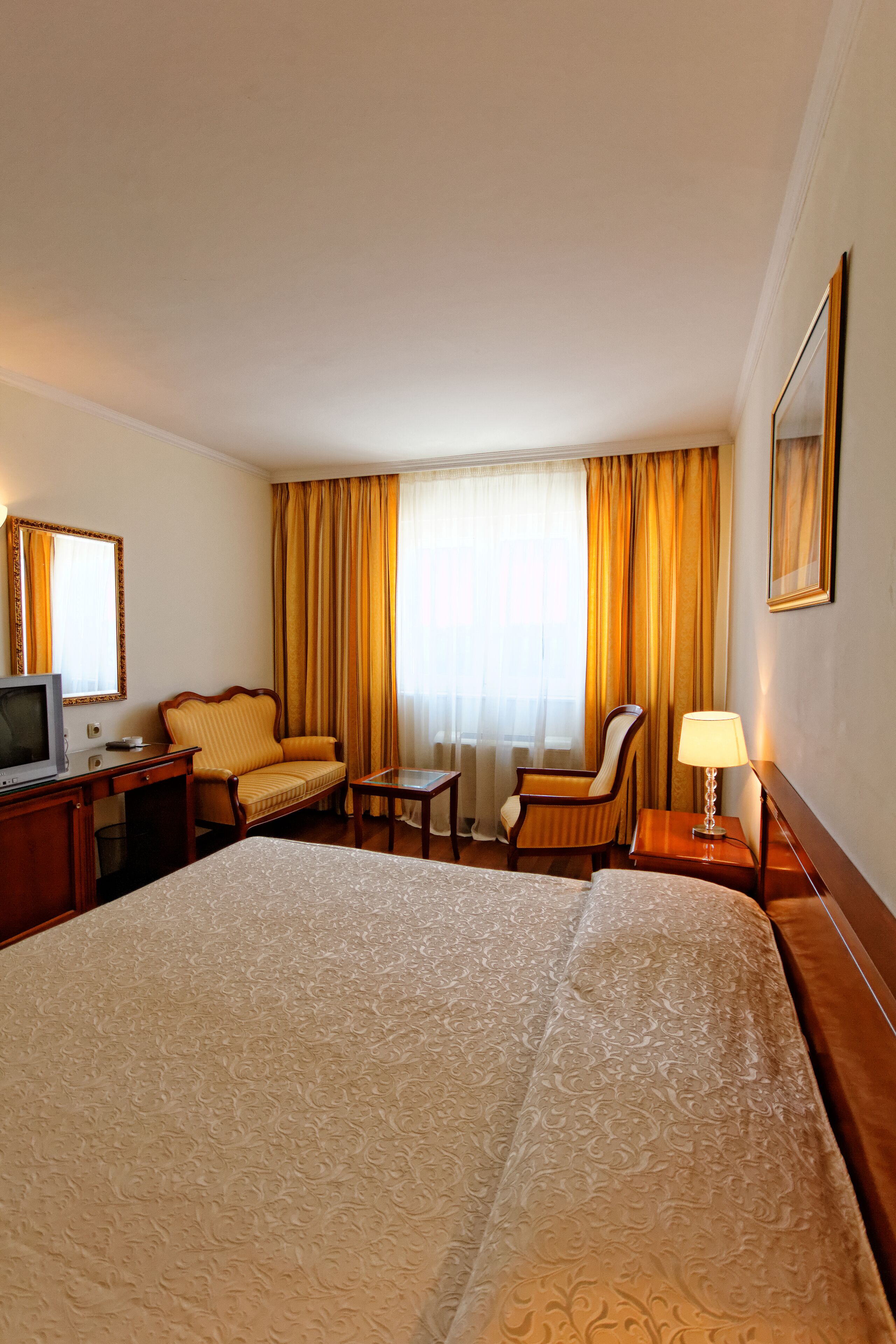 A hotel room with a large bed, a desk with a television, two armchairs, a small table, and a lamp.
