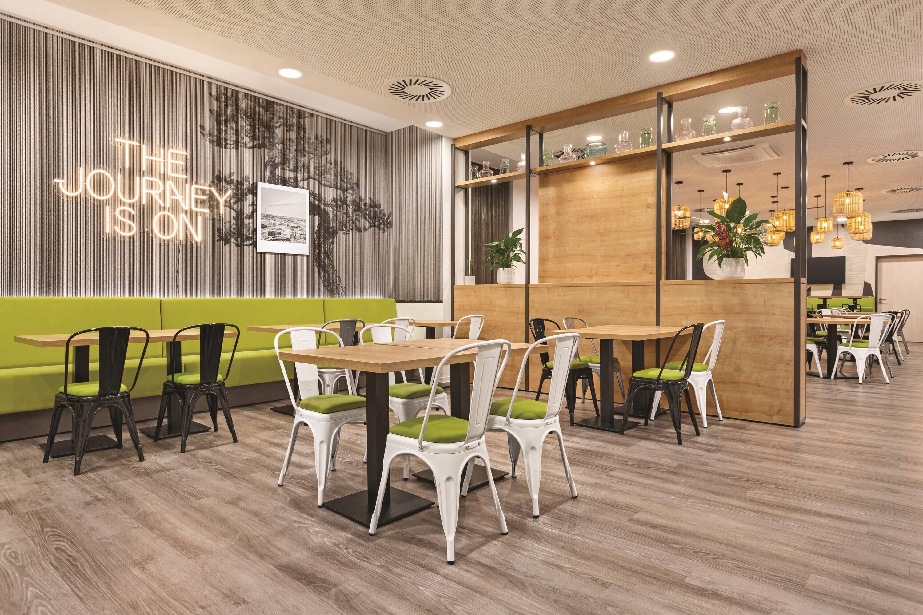 Ein modernes Restaurantinterieur mit Holztischen und -stühlen, einem grünen Sofa und einer Wand mit dem Schriftzug 'THE JOURNEY IS ON'.
