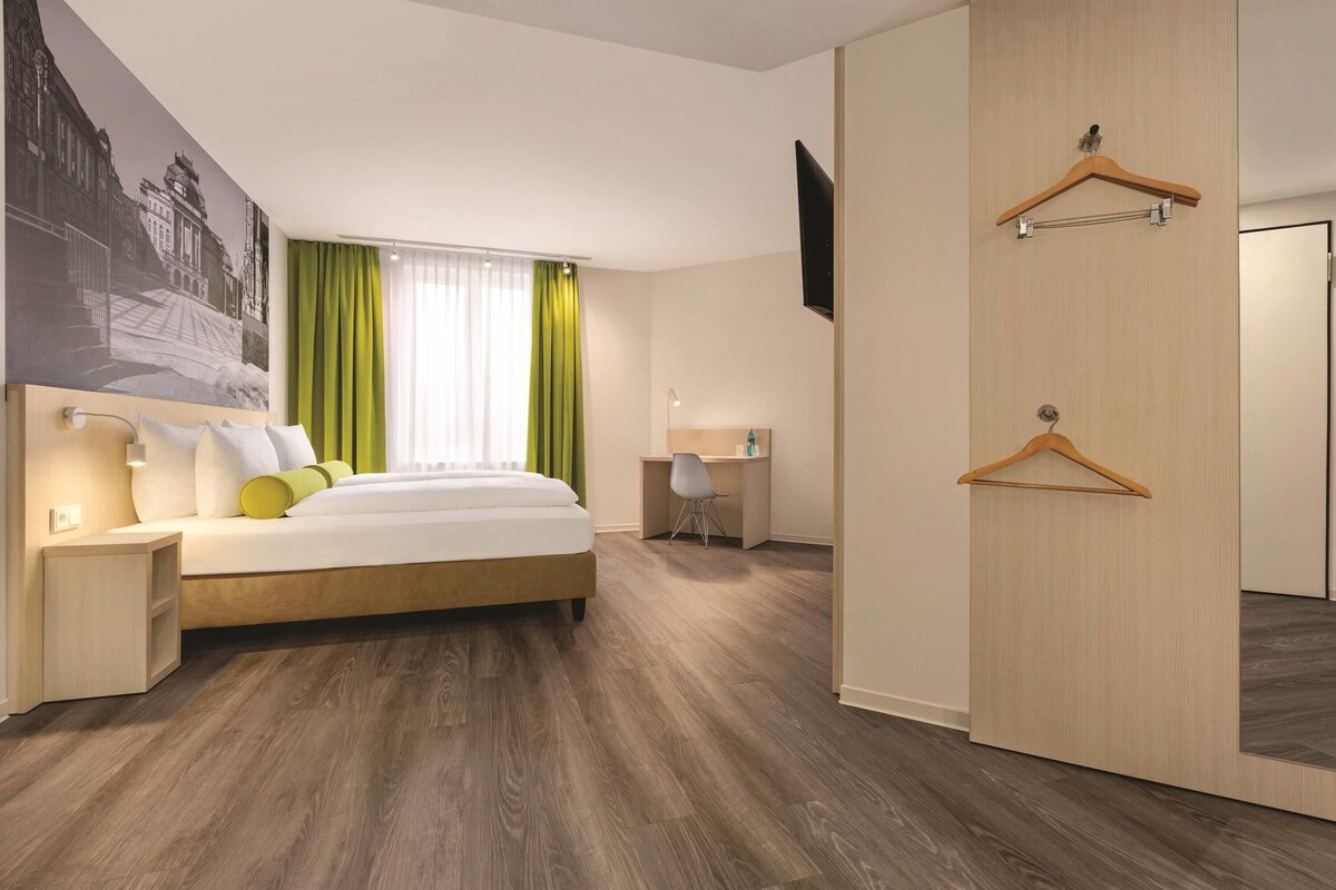 A modern hotel room with a large bed, a desk, and a wardrobe.