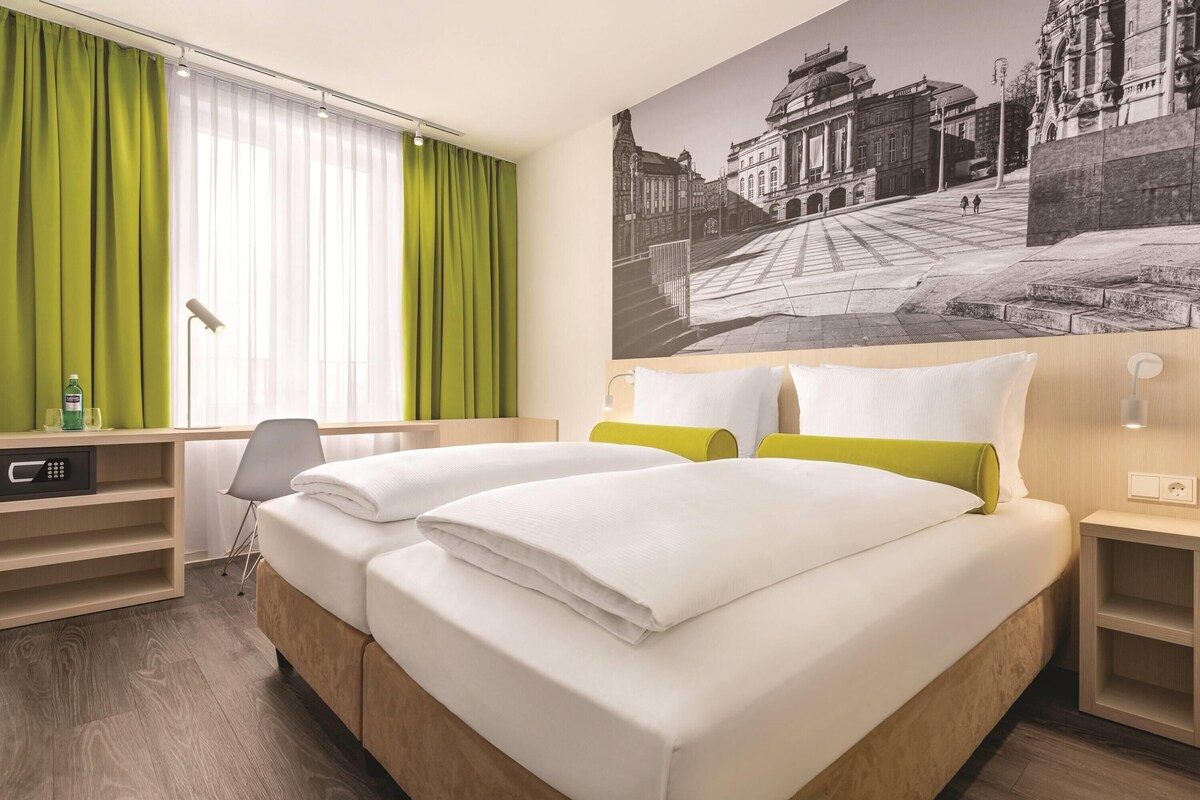 A hotel room with a bed, a desk, a chair, a window with curtains, and a wall-mounted lamp.
