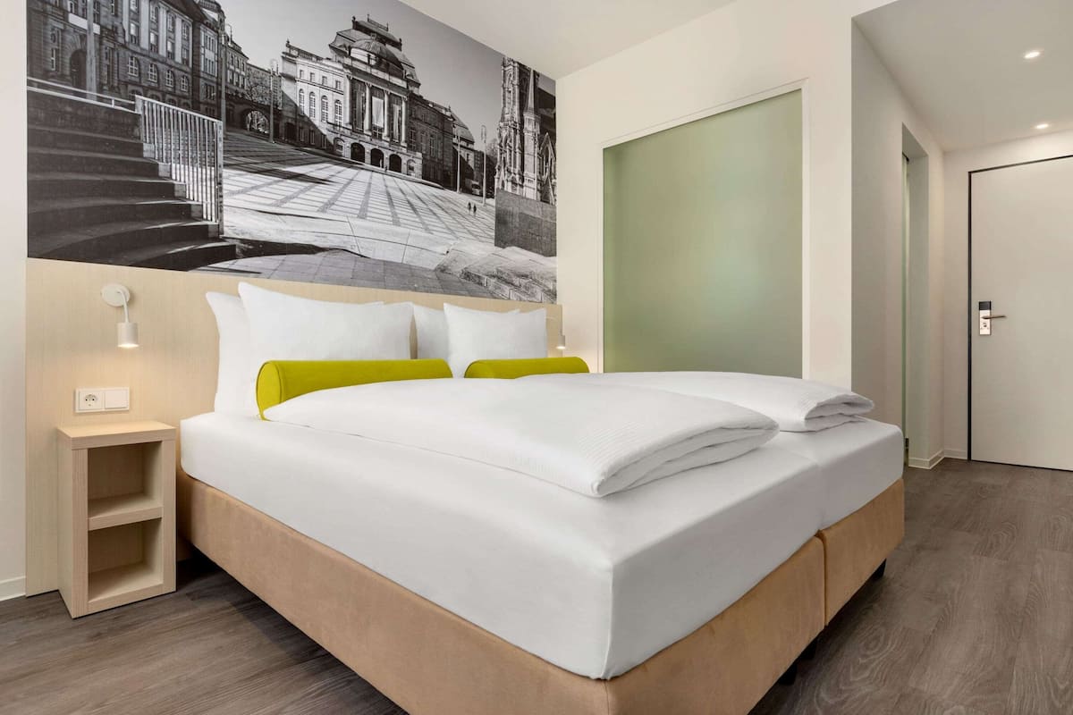 Une chambre d’hôtel moderne avec un grand lit, une table de chevet, une lampe fixée au mur et une grande fenêtre.