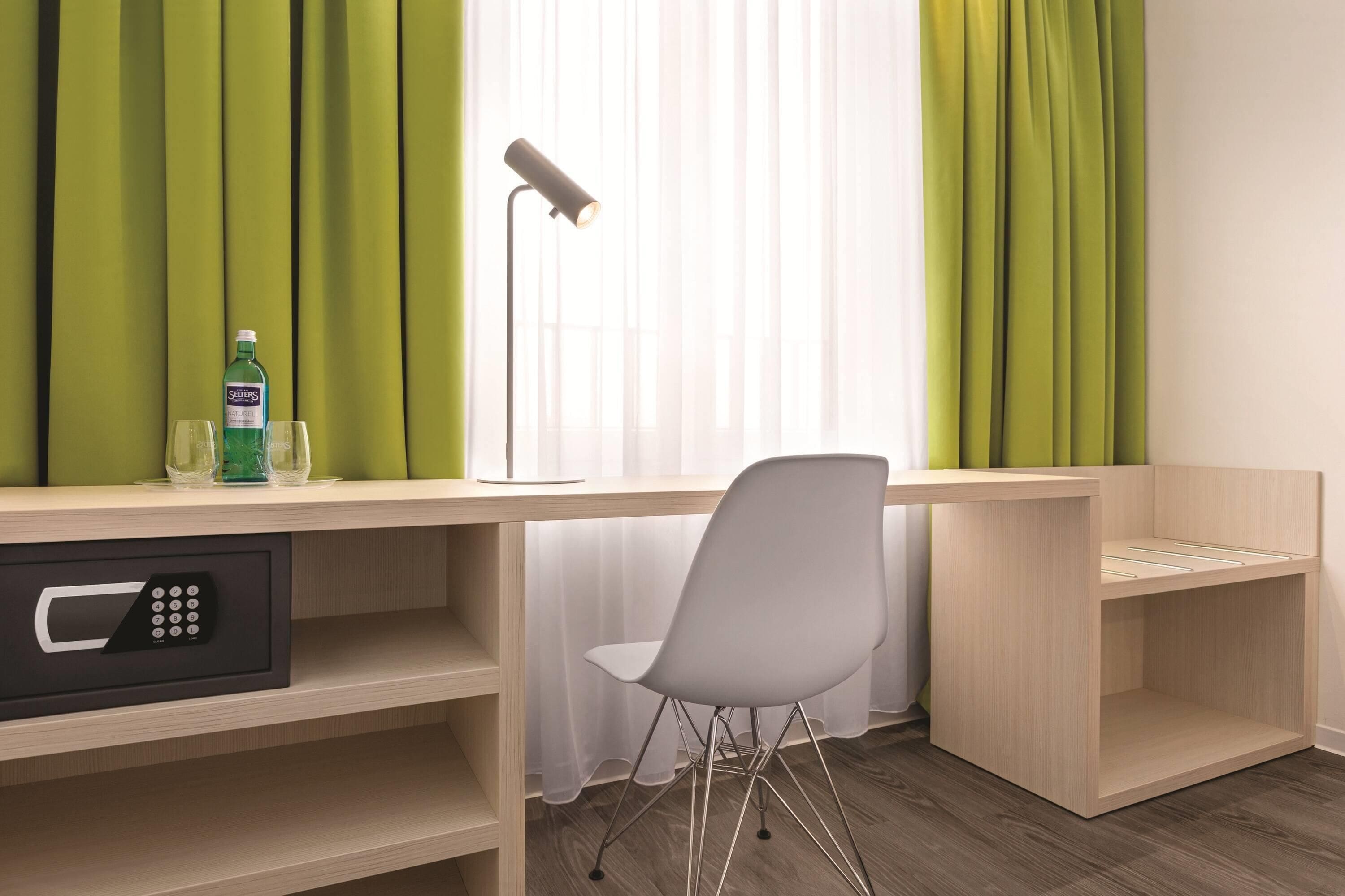 Ein Hotelzimmer mit Schreibtisch, Stuhl, Minibar und einem Fenster mit Vorhängen.