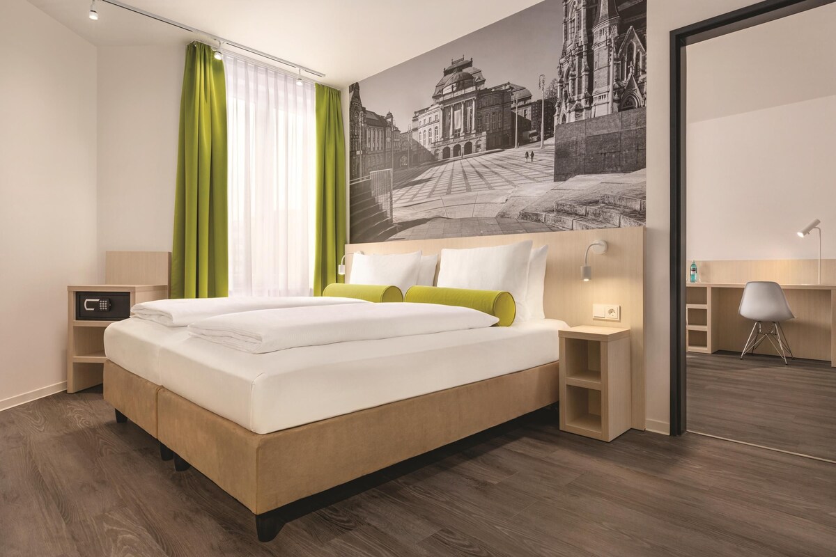A modern hotel room with a large bed, a desk, and a view of a historic building through the window.