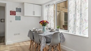 Un comedor con una mesa dispuesta para cuatro personas, una ventana con cortinas florales y un armario blanco sobre la mesa.