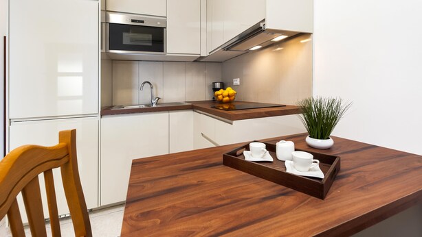 Una cocina moderna con armarios blancos, un horno de acero inoxidable, una mesa de comedor de madera con una bandeja de tazas y platitos blancos, y una planta en maceta.