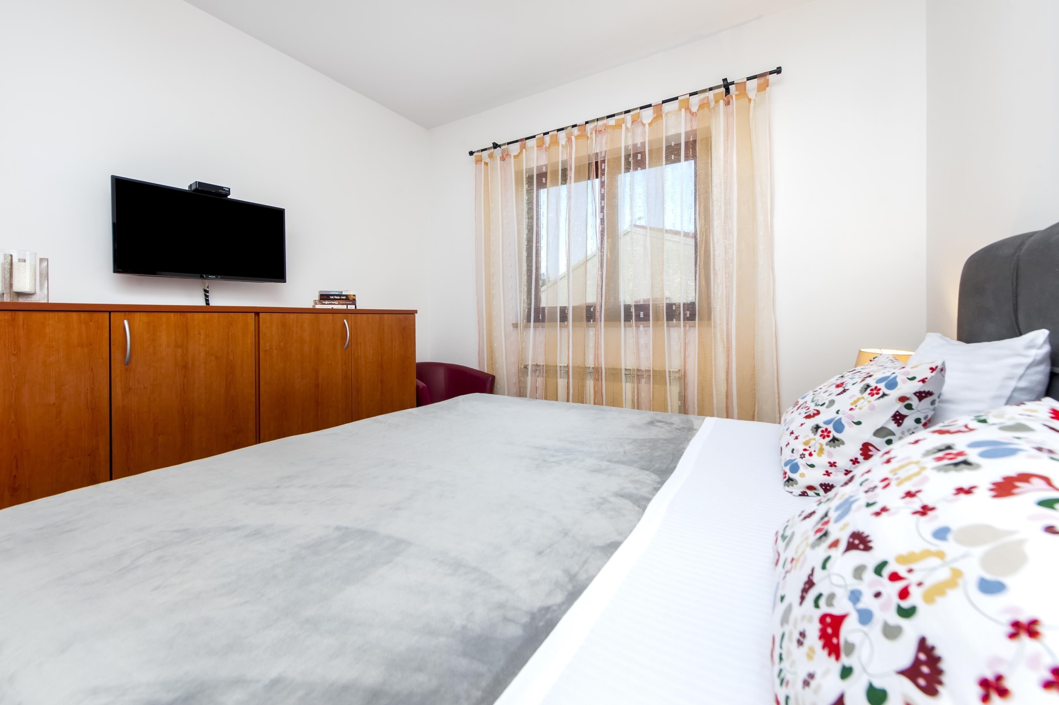 A bedroom with a bed, a TV mounted on the wall, a wooden wardrobe, and a window with sheer curtains.