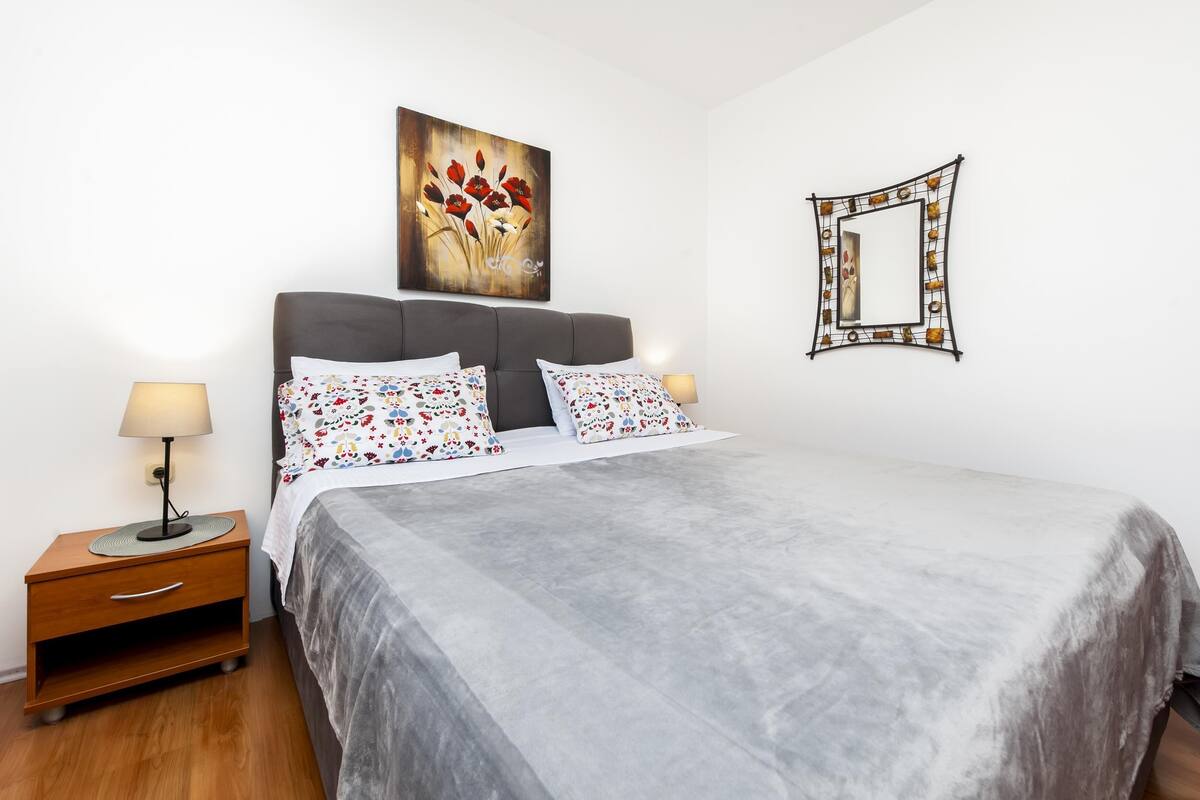 A neatly made bed with a grey bedspread and floral pillows, a bedside table with a lamp, a wall-mounted mirror, and a painting of red flowers.