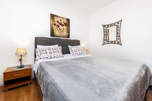 A neatly made bed with a grey bedspread and floral pillows, a bedside table with a lamp, a wall-mounted mirror, and a painting of red flowers.