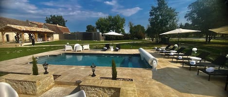 Una piscina amb gandules i ombrel·les, envoltada d'un paviment de pedra i una pèrgola de fusta.