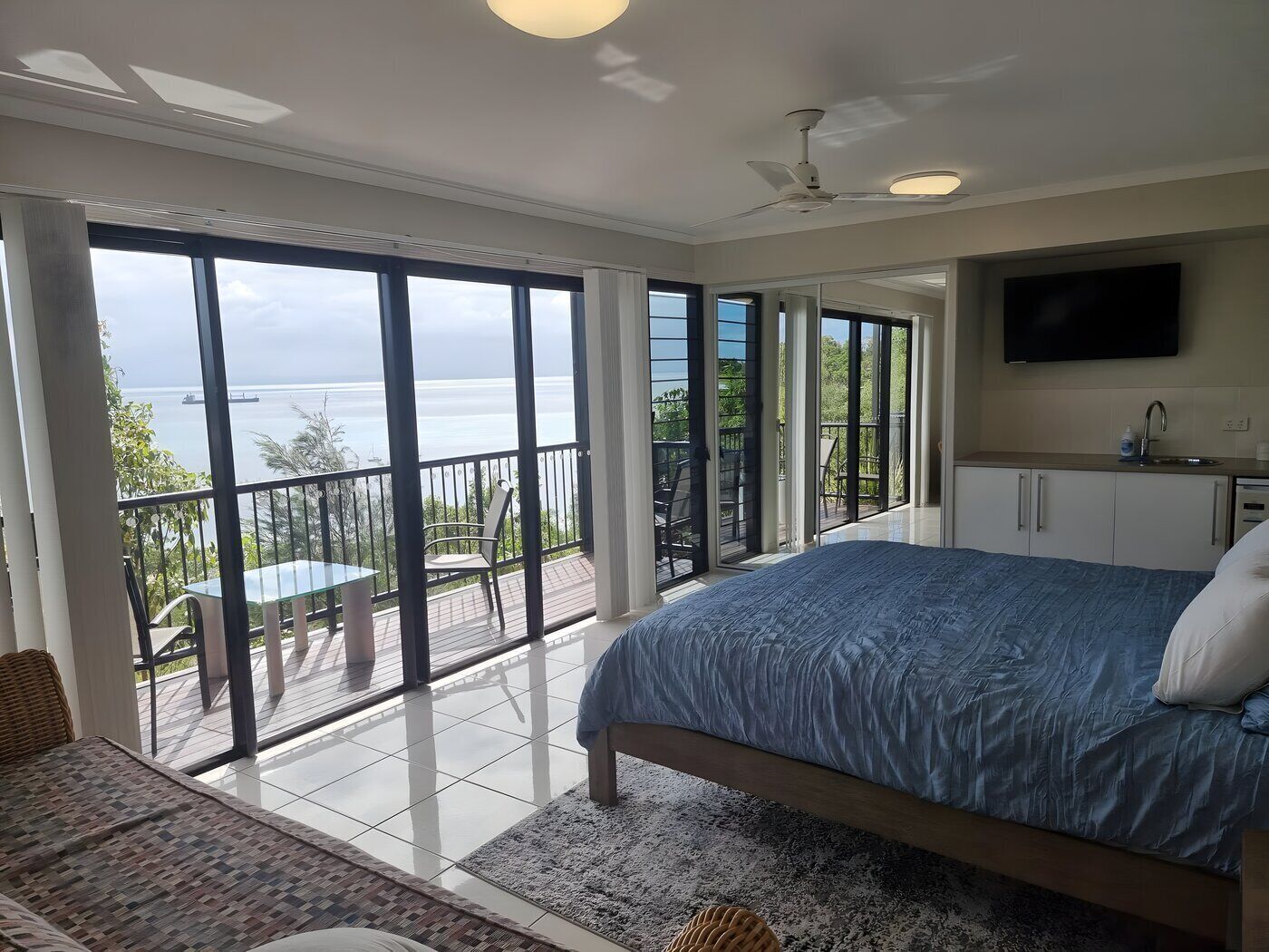 A bedroom with a large bed, a television, a balcony with a view of the sea, and a small kitchenette.