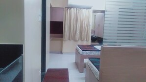 A hallway with a bed, a small table, and a mirror. There is a window with blinds and a curtain. - Choice Service Apartment (Pune)
