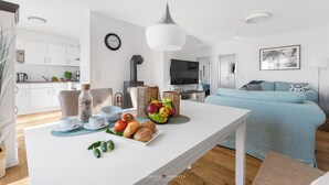 Een moderne keuken met witte kasten, een eettafel met ontbijtartikelen, een blauwe bank en een televisie op de achtergrond.