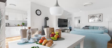 A modern kitchen with white cabinets, a dining table with breakfast items, a blue sofa, and a TV in the background.