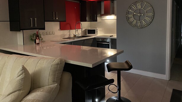 A modern kitchen with dark cabinets, a central island, and a large wall clock. - Cumberland stable lodge  (Workington)