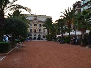 Una plaza abierta con palmeras, bancos y un edificio histórico con banderas.