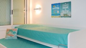 A single bed with a teal bedspread in a room with a window and a wall-mounted poster.