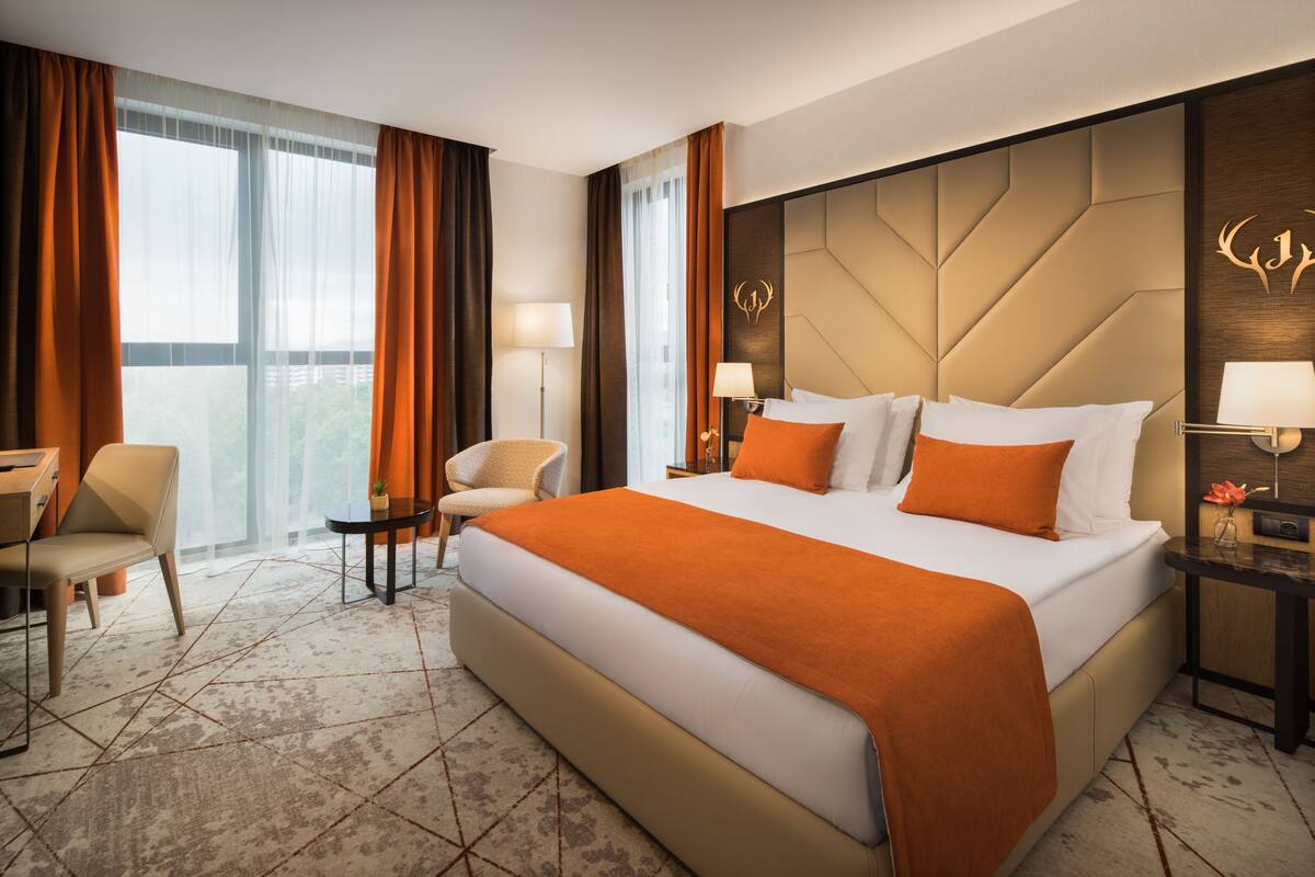 Une chambre d’hôtel avec un grand lit, deux tables de chevet avec des lampes, un fauteuil et une vue sur la ville grâce à de grandes fenêtres.