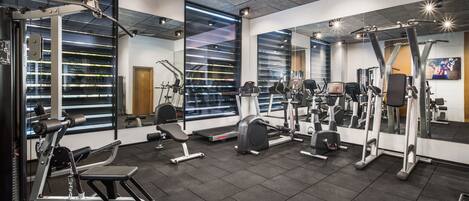 Ein modernes Fitnessstudio mit verschiedenen Trainingsgeräten, großen Fenstern mit Jalousien und einer Spiegelwand.