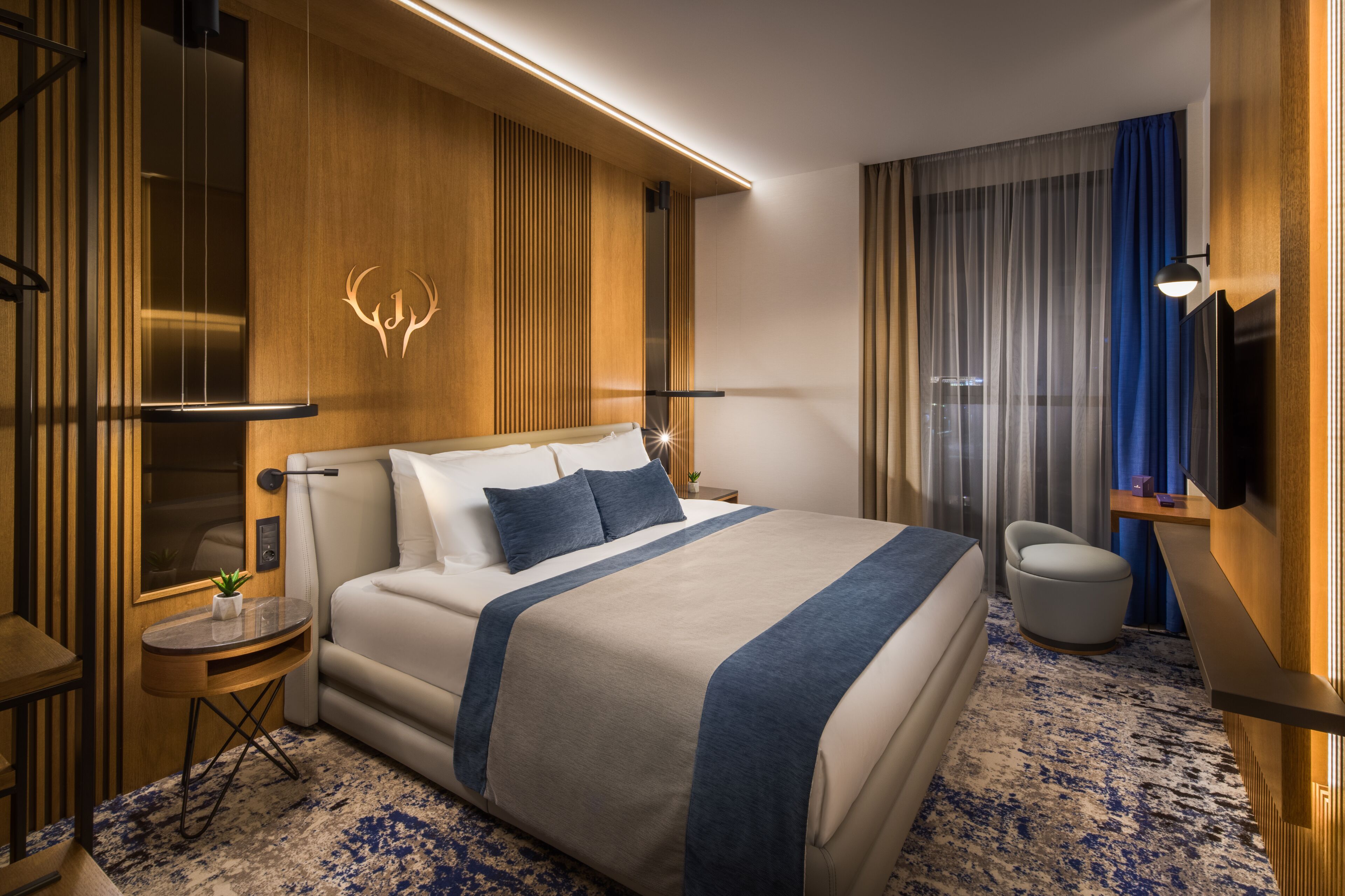A hotel room with a large bed, a small side table, a chair, a TV, and a wooden accent wall with a light fixture.