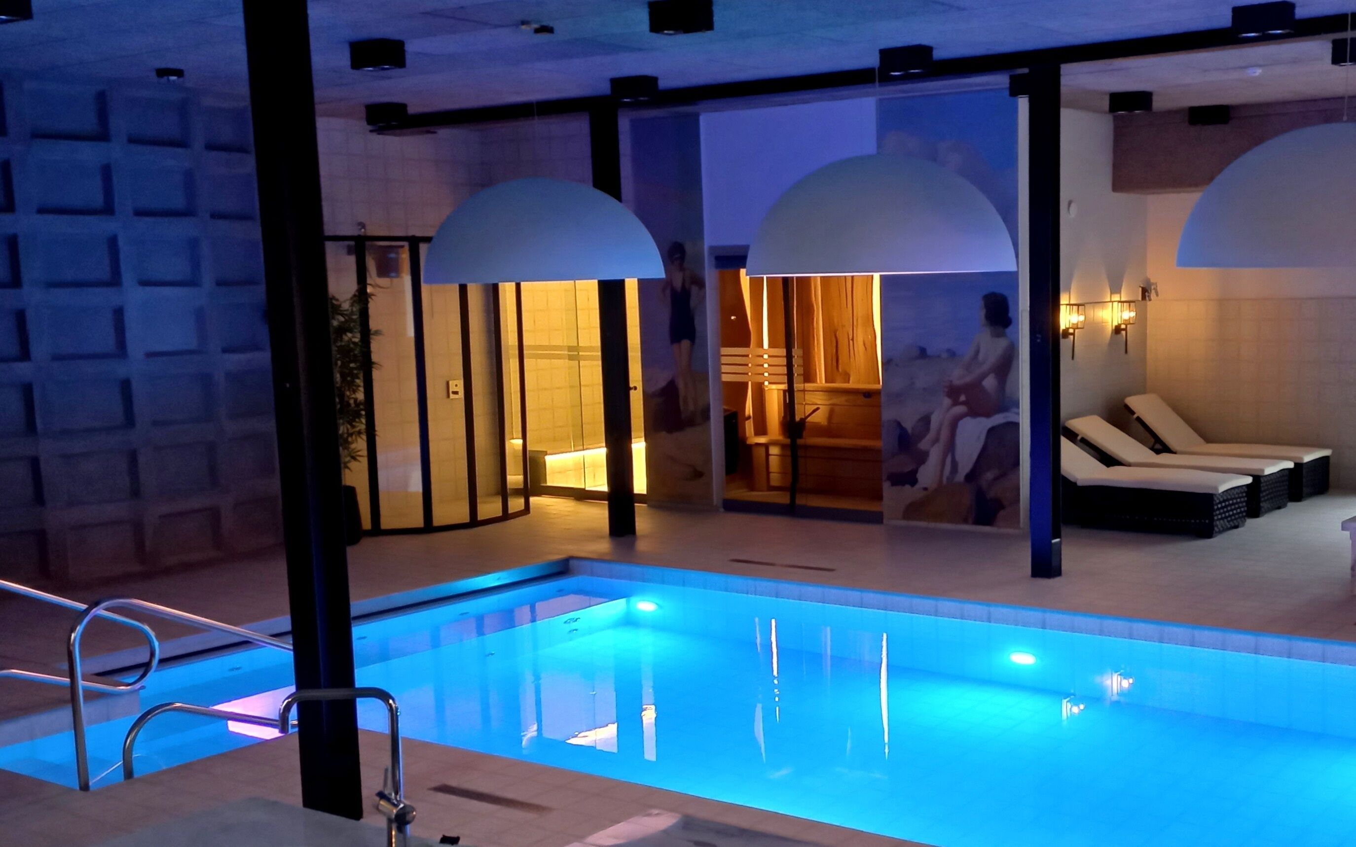 A modern indoor pool area with a clear pool, lounge chairs, and a glass partition separating the pool from the adjacent room.