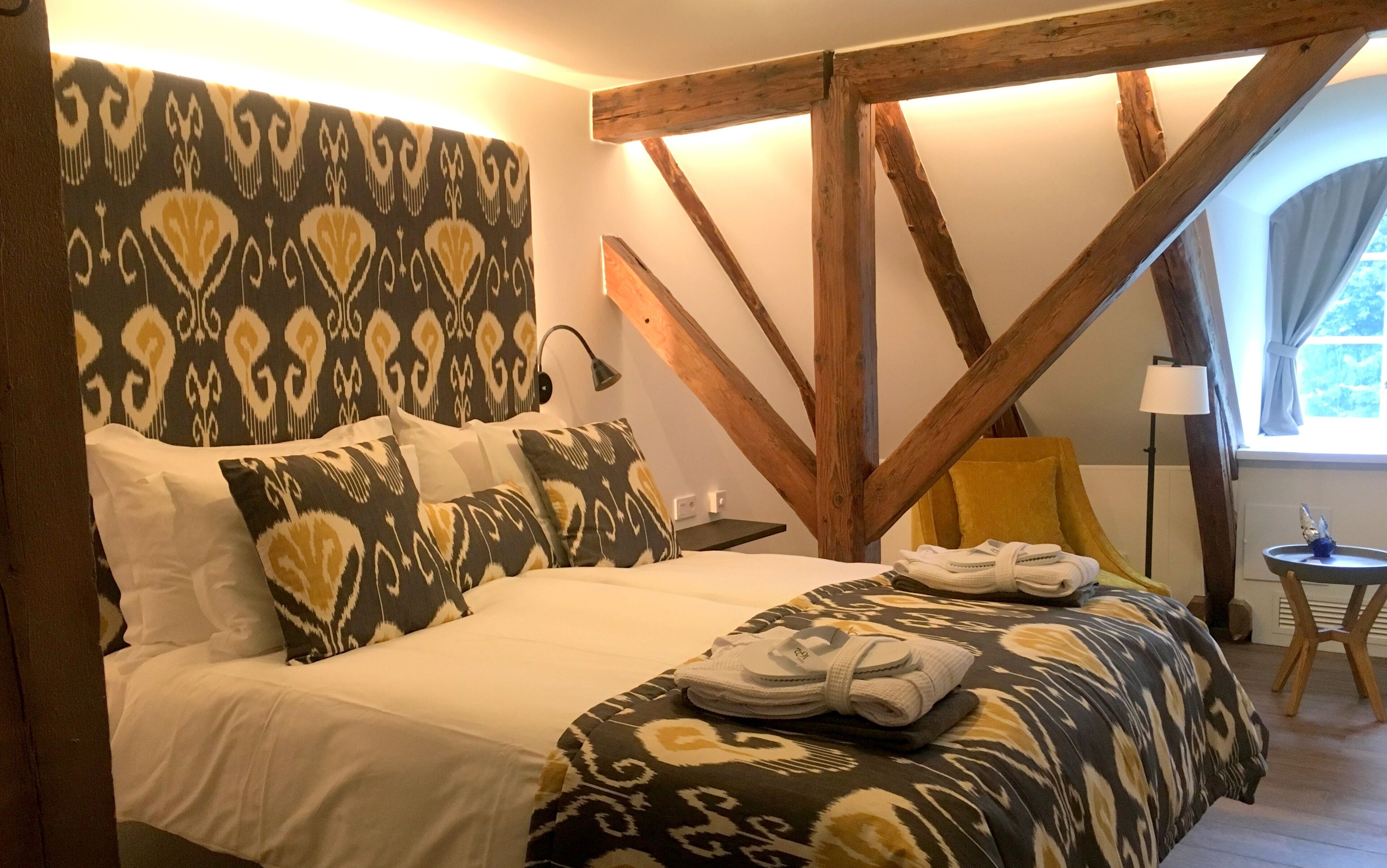 A bedroom with a bed, patterned headboard, wooden beams, a small table, and a chair.