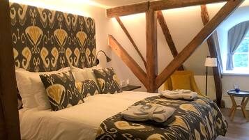 A bedroom with a bed, patterned headboard, wooden beams, a small table, and a chair.