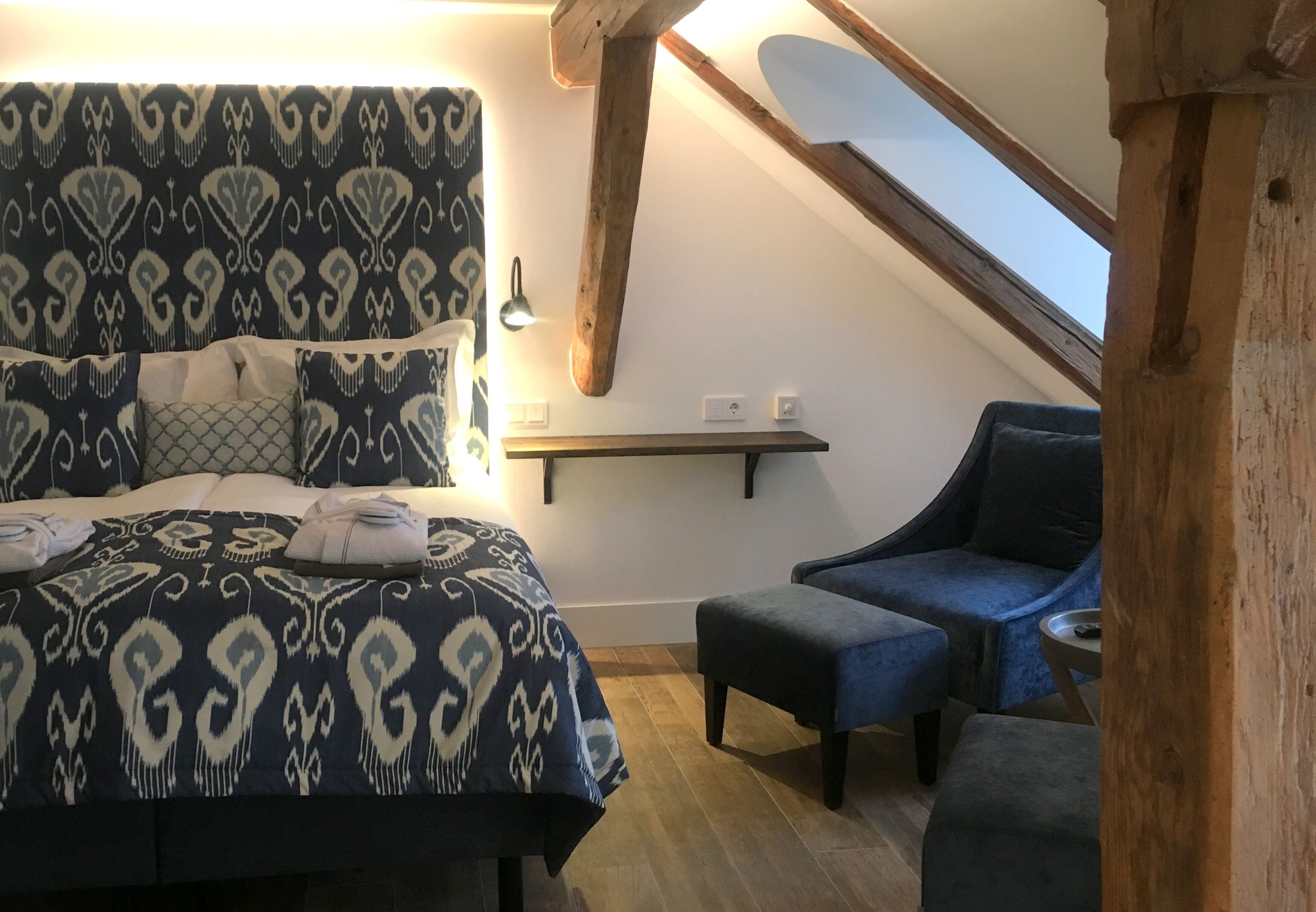 Une chambre avec une couvre-lit à motifs, une poutre en bois au plafond, une petite table et une chaise bleue.