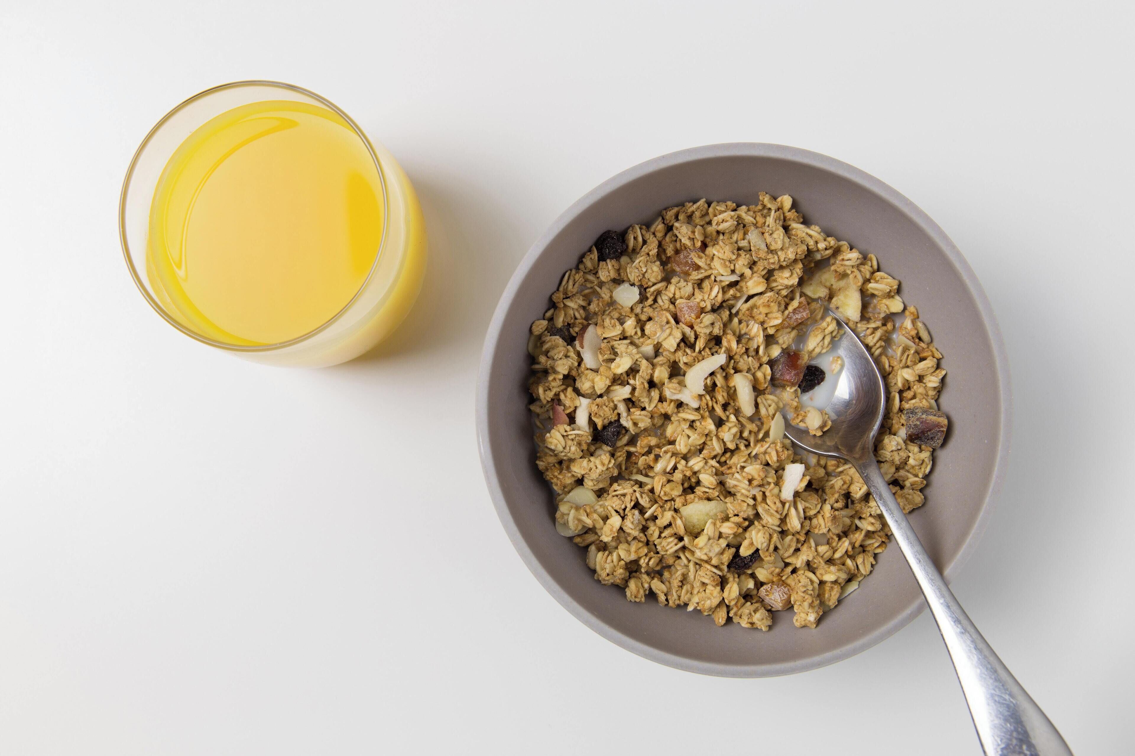 A bowl of granola with a spoon and a glass of orange juice on a white surface.