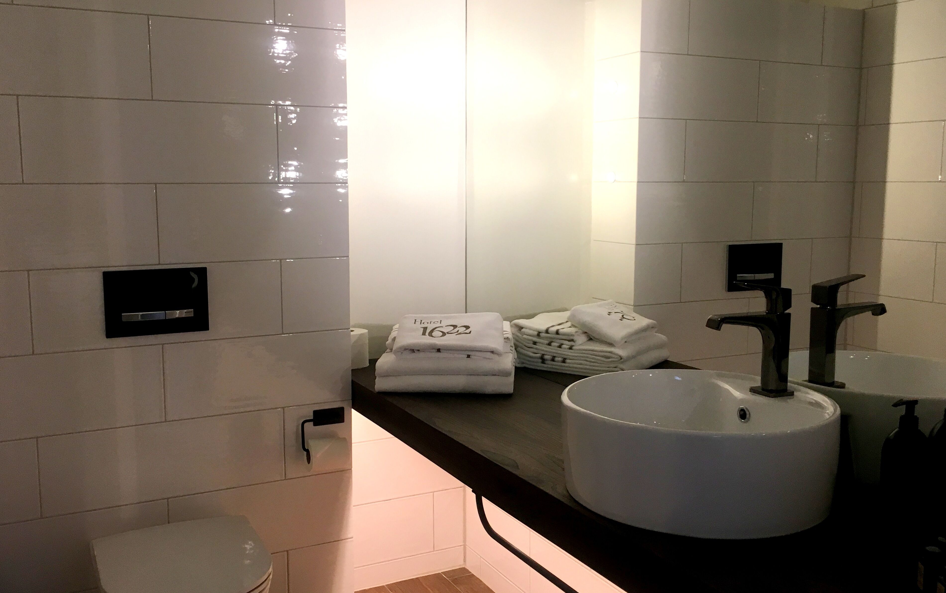A modern bathroom with white tiles, a round sink, two black faucets, and a stack of white towels with a hotel logo.
