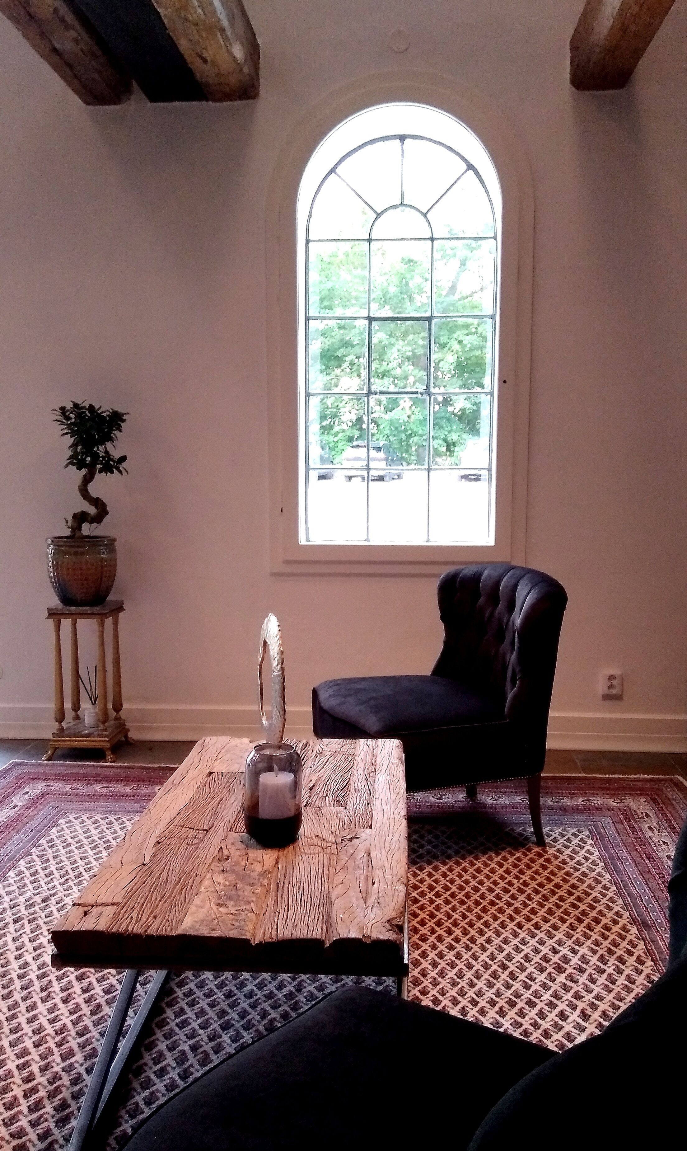 Una habitación con un amplio ventanal arqueado, una planta en maceta, una silla, una mesa de centro de madera y un tapete estampado.