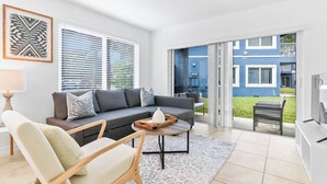 Uma sala de estar moderna com um sofá cinzento, uma mesa de centro de madeira e uma poltrona bege. Há uma televisão à direita e uma janela com persianas à esquerda.
