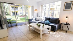 A modern living room with a sofa, coffee table, and a view of a garden through large windows. - 320B - Boca Retreat w/ Pool, Parking, Near Evert Tennis & Shops (Boca Raton)