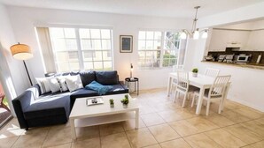 A modern living room with a sofa, coffee table, and dining area. There are large windows providing natural light and a view of the outside. - 320B - Boca Retreat w/ Pool, Parking, Near Evert Tennis & Shops (Boca Raton)
