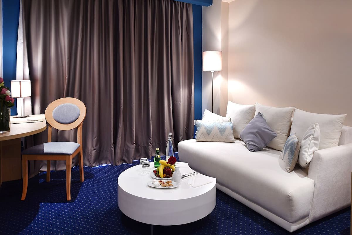 A hotel room with a round white coffee table, a white sofa with multiple pillows, a wooden chair, a dining table with a lamp, and a large window with curtains.