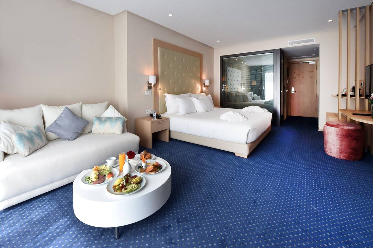 A hotel room with a bed, a sofa, a round coffee table with breakfast, and a large window.