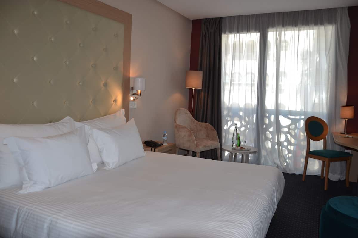 A hotel room with a large bed, a chair, a desk, and a window with curtains.