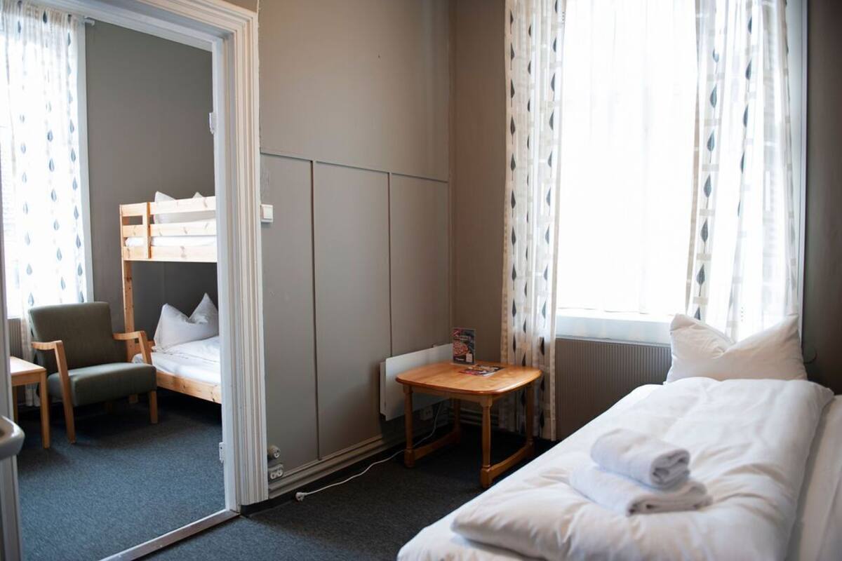 A compact hotel room with a bunk bed, a single bed, a small table, a chair, and a radiator.