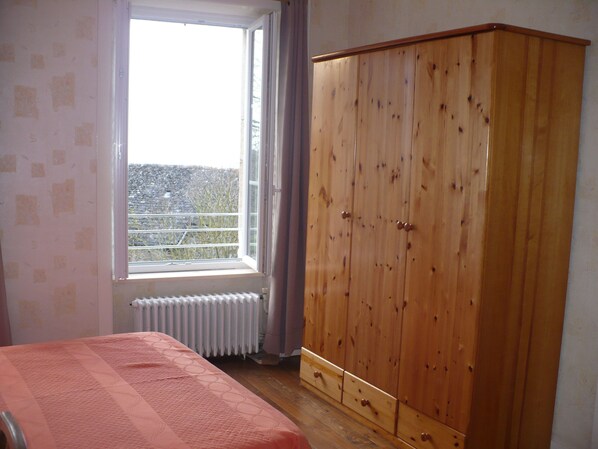 Un dormitorio con un armario de madera, un radiador y una ventana con vistas a una valla y árboles.