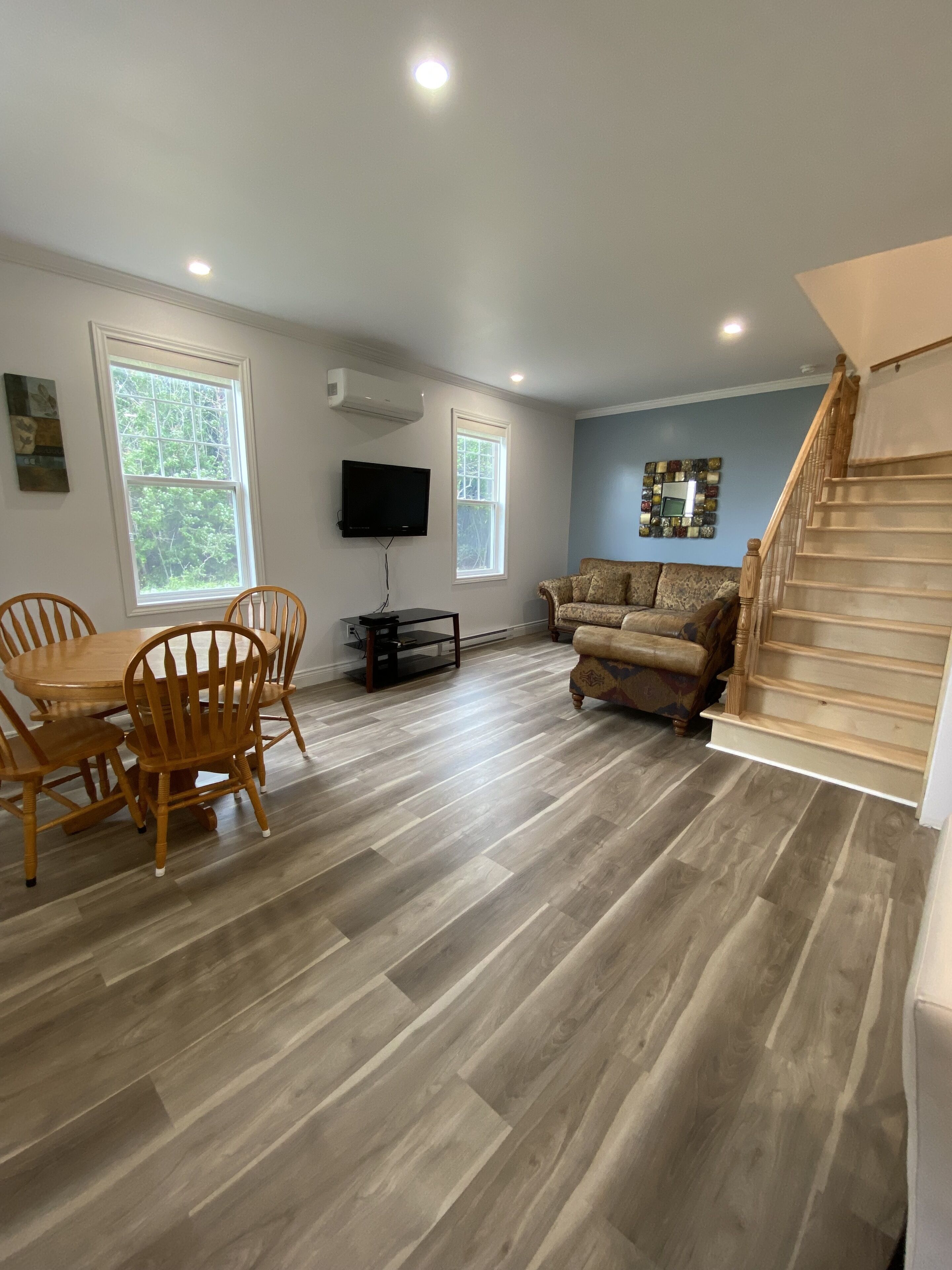 Un vaste salon comprenant une table et des chaises en bois, un canapé, un téléviseur fixé au mur et un escalier menant à un autre niveau.